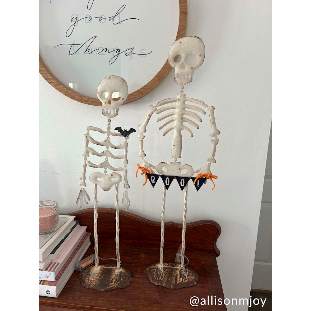 Two decorative Halloween skeleton figurines on a wooden table next to stacked books and a wall sign.