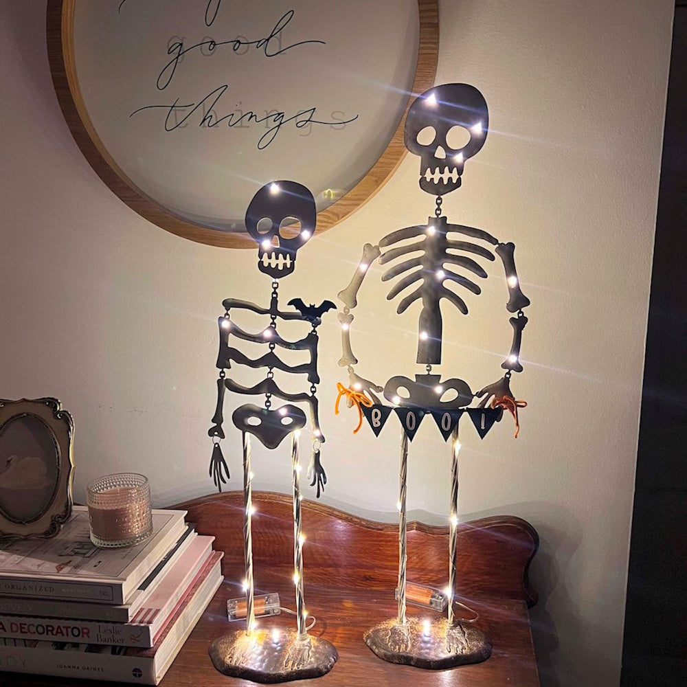 Two light-up metal Halloween skeletons glowing on a wooden table next to stacked books and a candle.