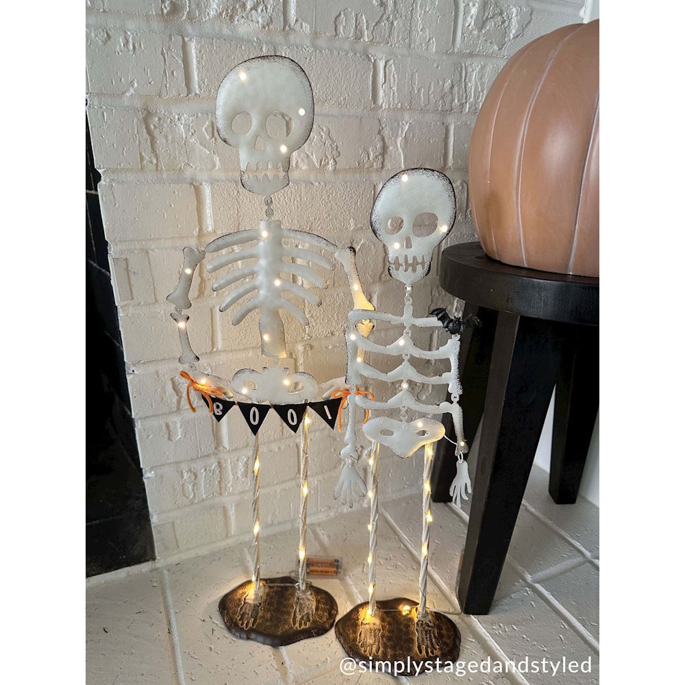 Two light-up Halloween skeleton stakes holding a Boo banner next to a black wooden stool.