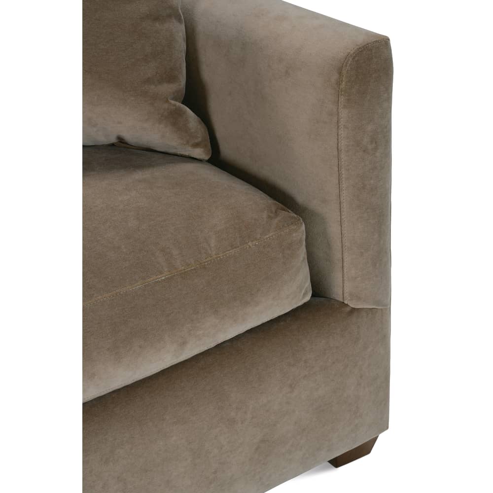 Close-up of a modern beige velvet sofa showing a plush seat cushion, a fabric armrest, and a wooden leg.