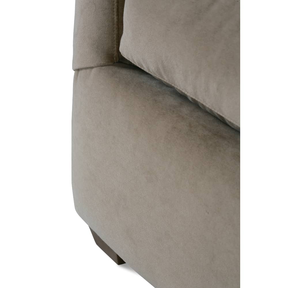Close-up of a taupe velvet sofa showing its plush cushion, upholstered corner, and short wooden leg.