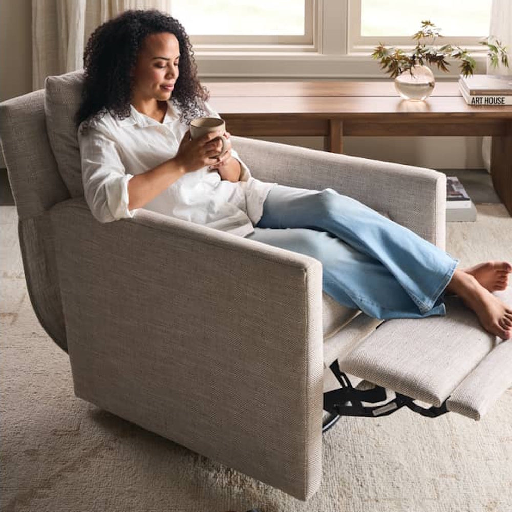 A woman relaxes in a beige upholstered power recliner while holding a mug in a modern, sunlit living room.