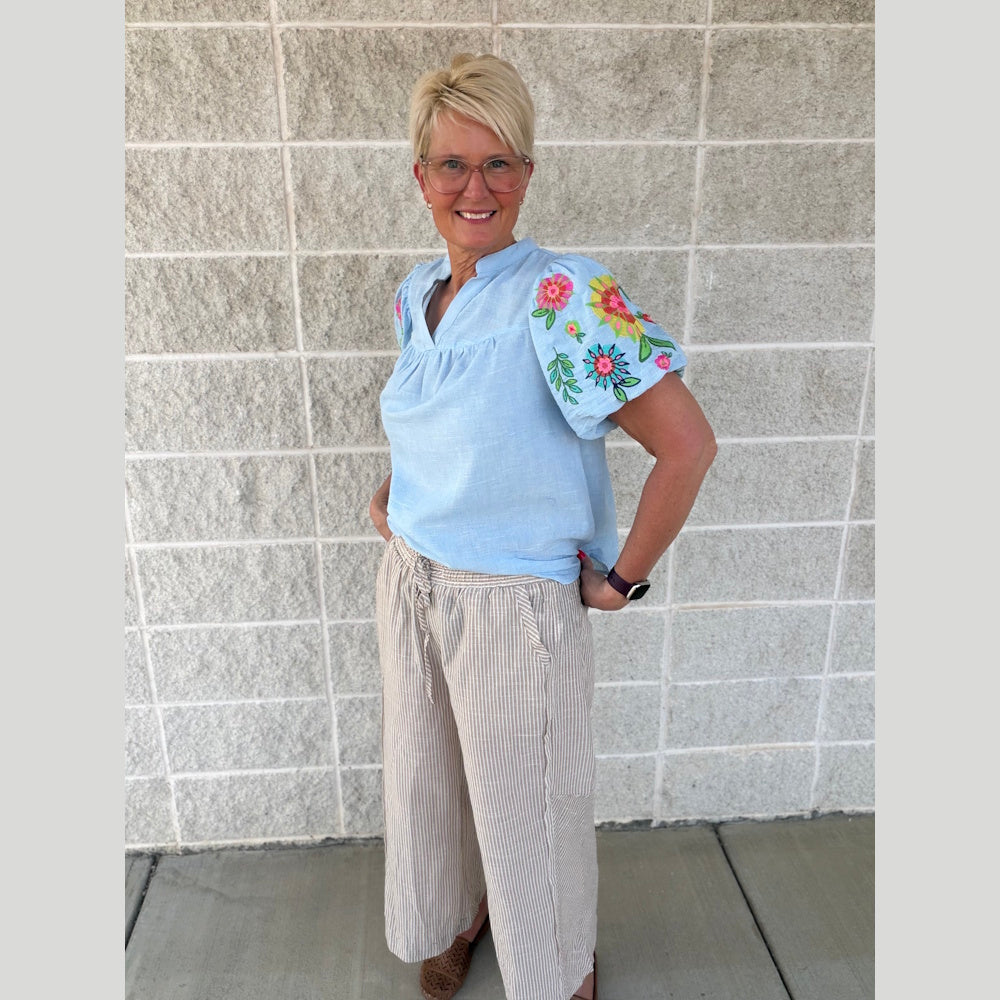 Woman in a light blue embroidered top and tan striped wide-leg linen pants posing against a brick wall.