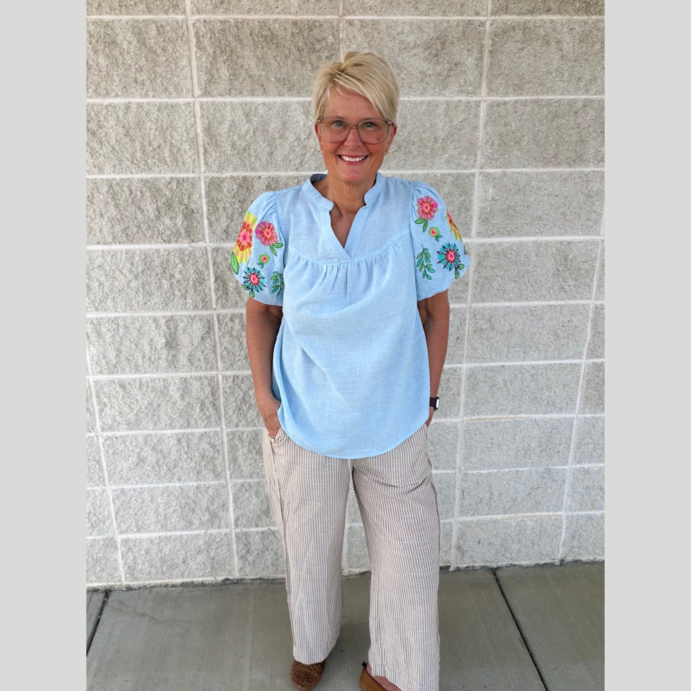 Woman in a light blue embroidered puff-sleeve top and tan striped wide-leg linen pants standing against a brick wall.