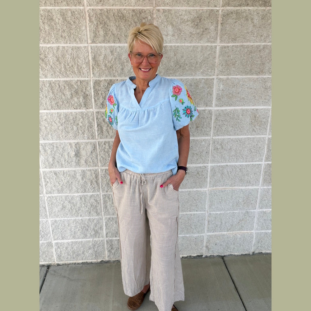 Smiling woman with short blonde hair wearing a light blue embroidered blouse and striped wide-leg linen pants.