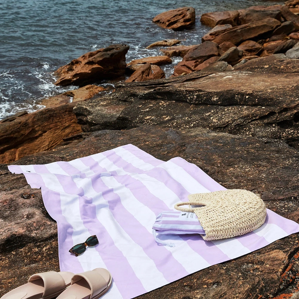 A lilac and white striped beach towel spread on rocky shores, with a straw bag, sunglasses, and sandals.
