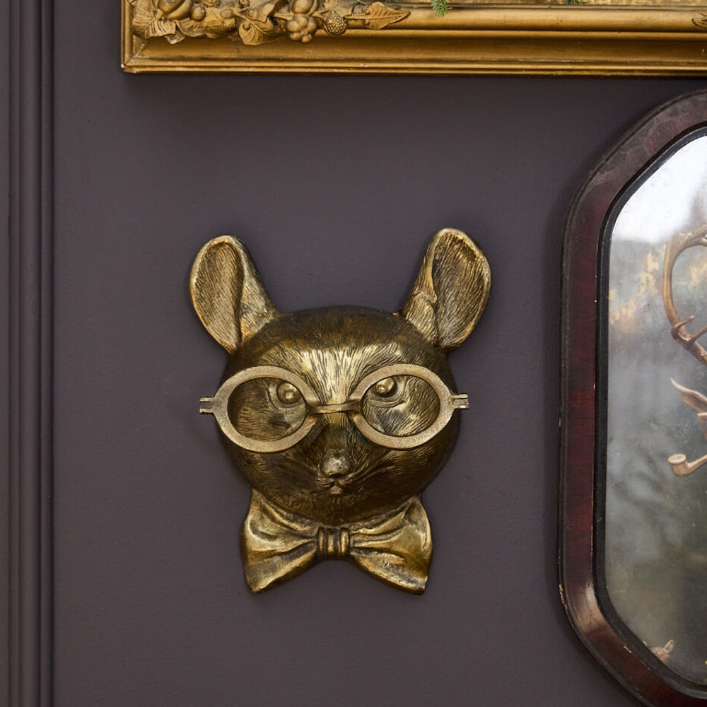A gold mouse head wall mount wearing round glasses and a bow tie, mounted on a dark purple wall.