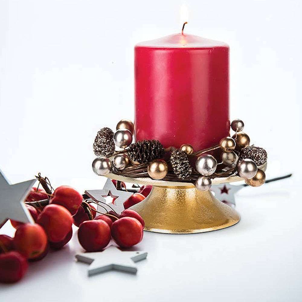 Lit red pillar candle on a gold pedestal stand, decorated with festive ornaments, berries, and stars.