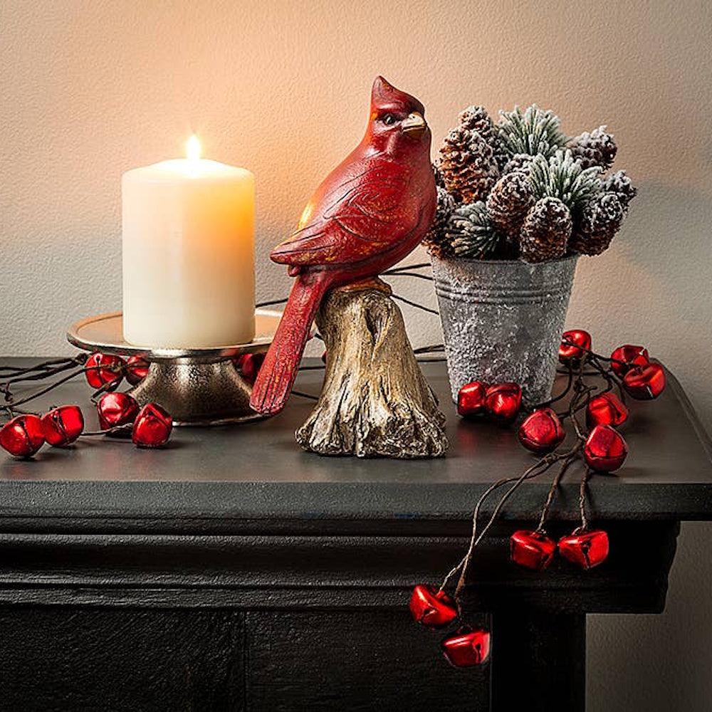Red cardinal figurine beside a lit candle on a stand, frosted pinecones, and red jingle bells.