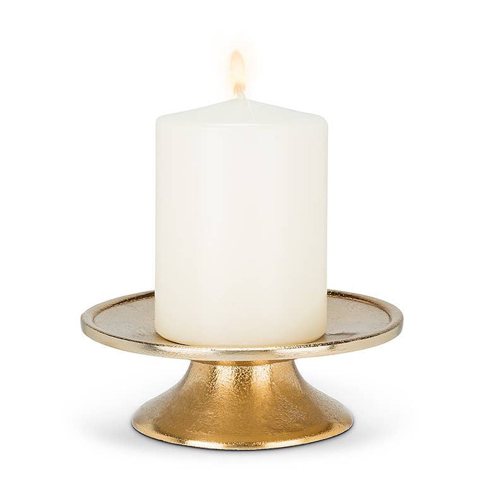Lit white pillar candle sitting on a textured gold pedestal plate on white background.