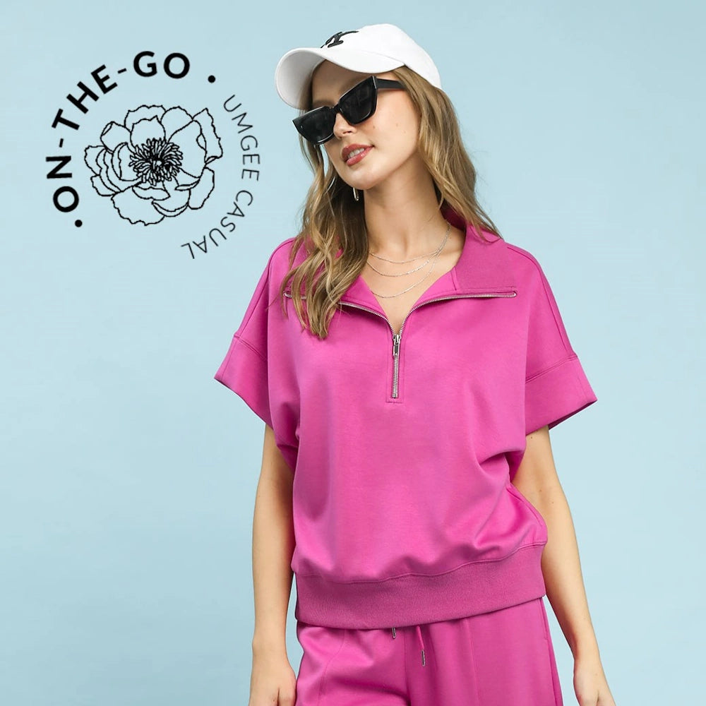 Woman in a pink Umgee short-sleeve half-zip pullover, white baseball cap, and sunglasses against a blue background.