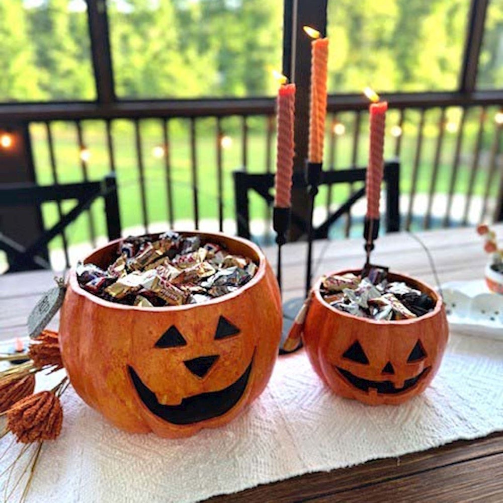 Two pumpkin candy bowls filled with Halloween treats on a table with lit orange candles.