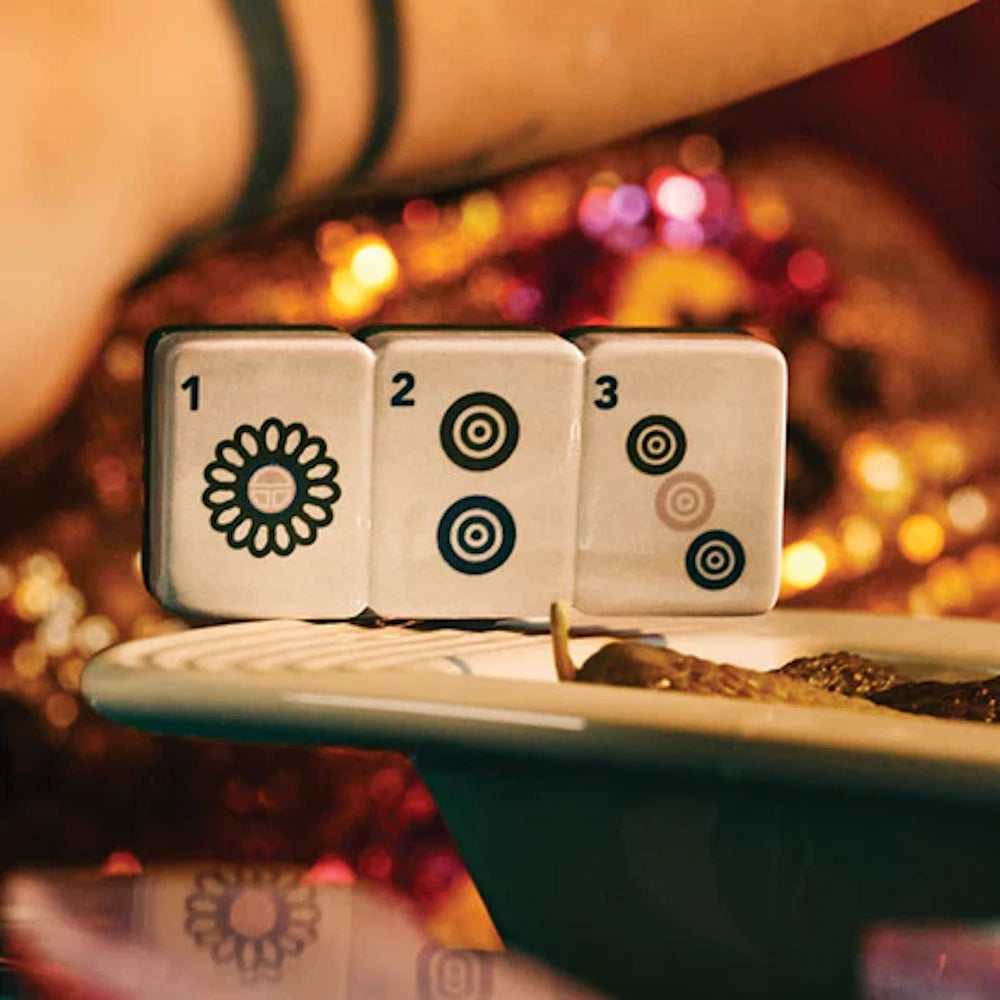 Three Mahjong tiles numbered 1, 2, and 3 resting on the edge of a ceramic tray against a bokeh background.