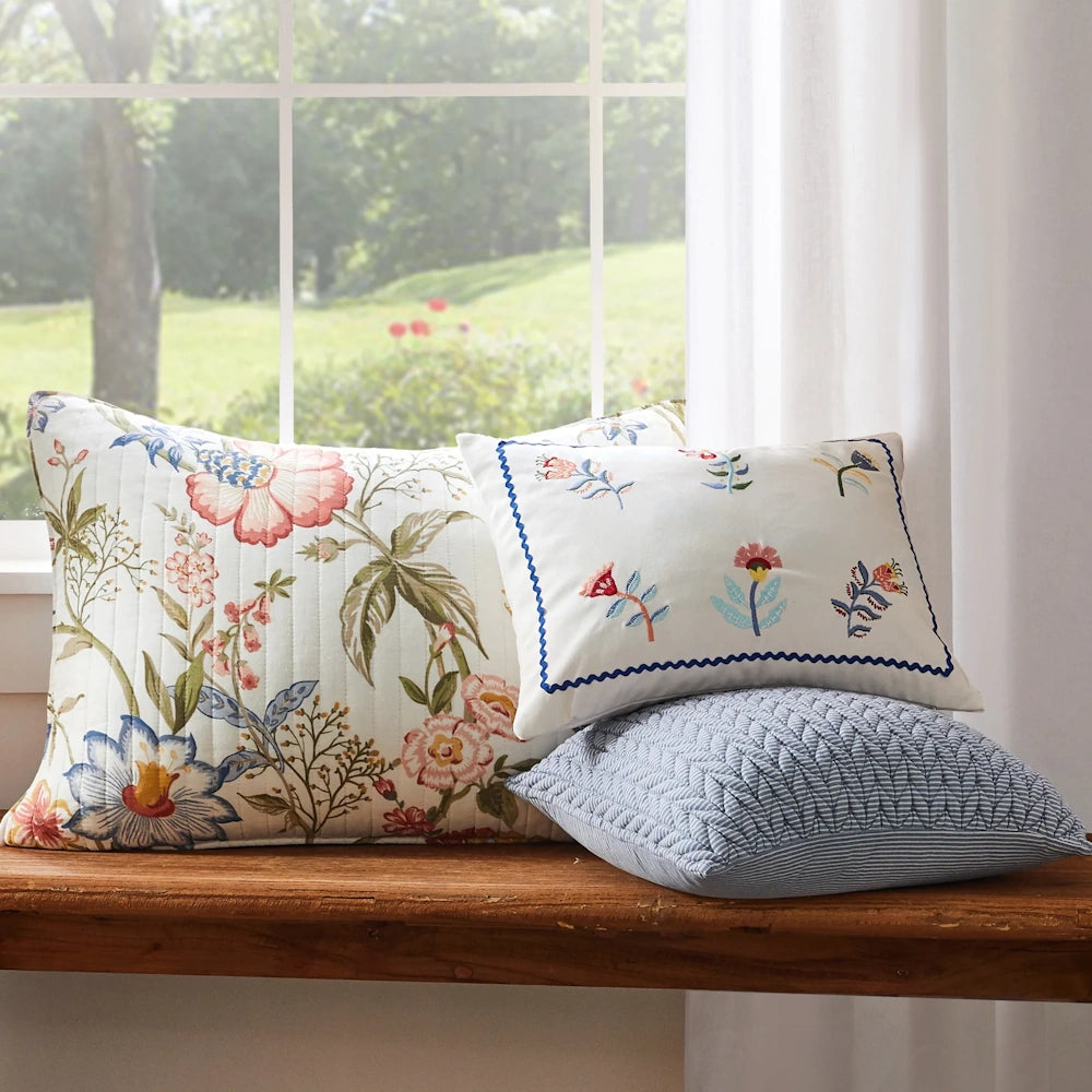 Floral pillow sham and decorative pillows resting on a wooden window seat next to a sunny window.