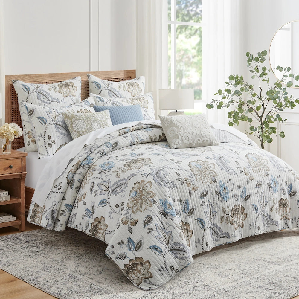 Neutral and blue floral quilt set styled on a bed with stacked pillows in a bright, modern bedroom.