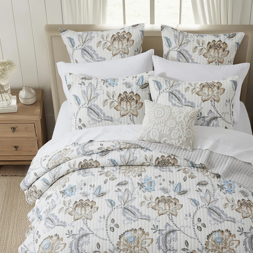 Neutral blue floral quilt set styled on a wooden woven bed with layered accent pillows in a bright bedroom.