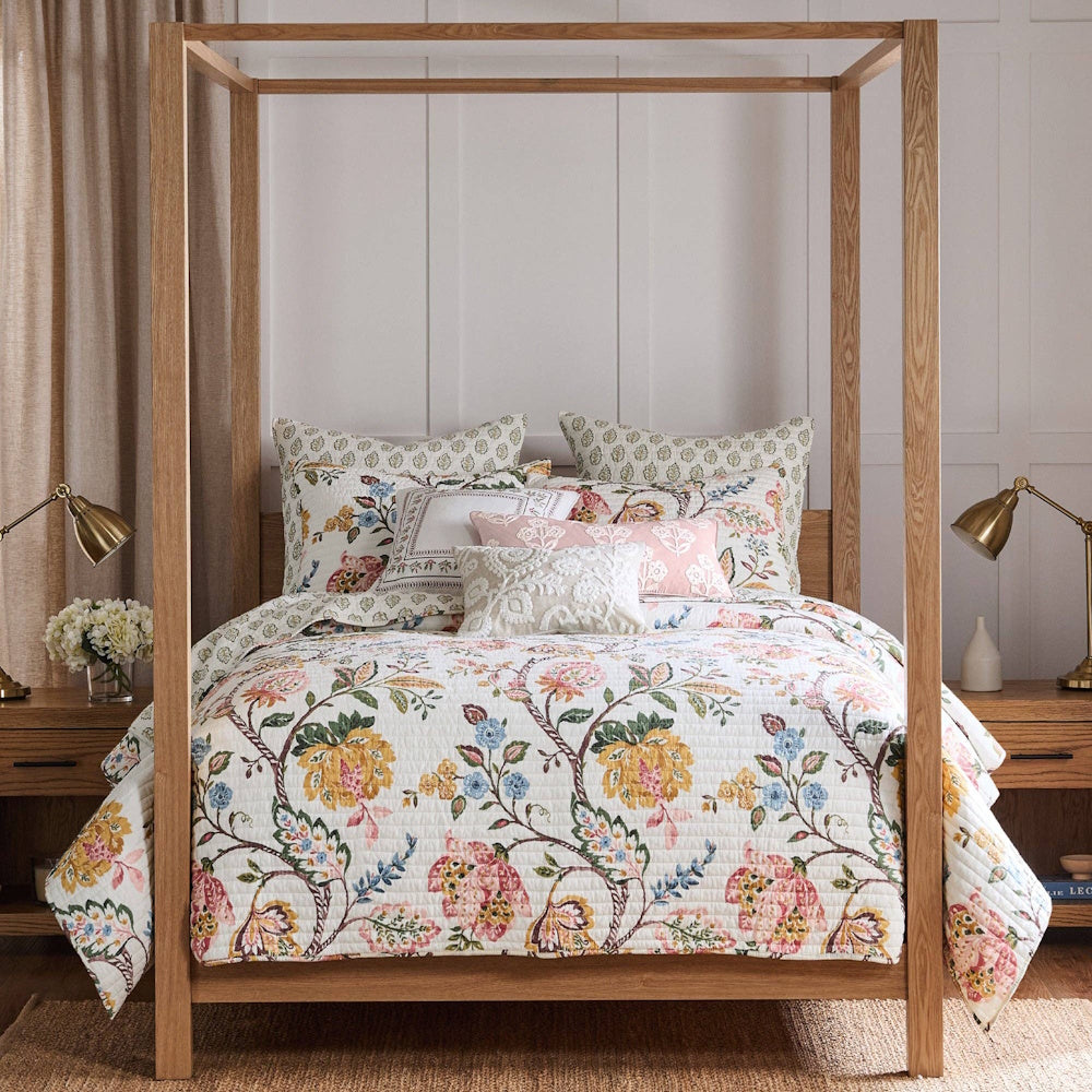Floral quilted bedding set on a wooden canopy bed with patterned pillows and gold lamps in a bright bedroom.