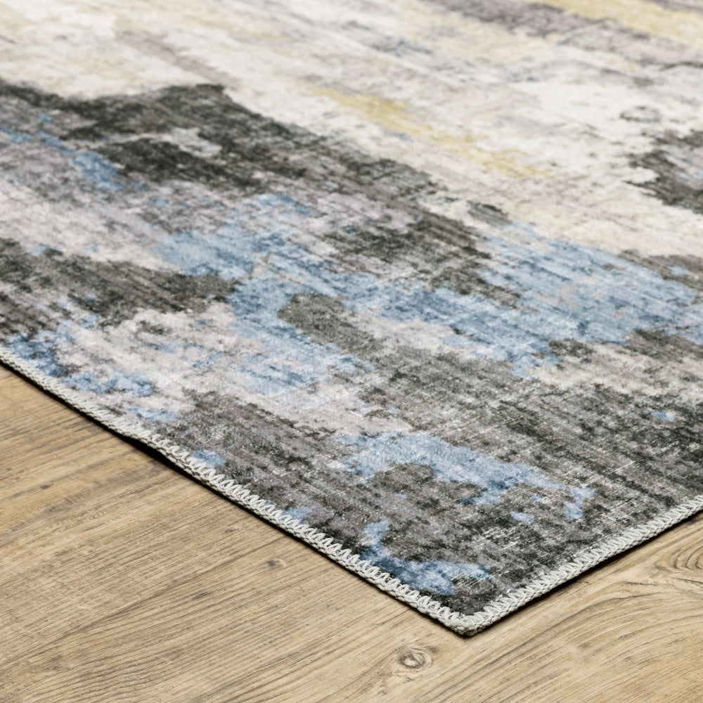 Close-up corner of an abstract area rug with mottled blue, grey, cream, and gold patterns on a wood floor.