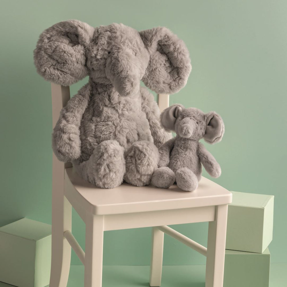 Mother and baby grey elephant plush toys sitting on a white wooden chair against a light green background.