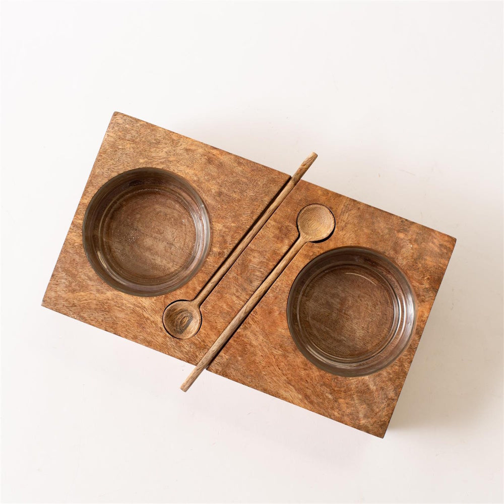 Top-down view of a wooden serving plank set with two glass bowls and two small wooden spoons.