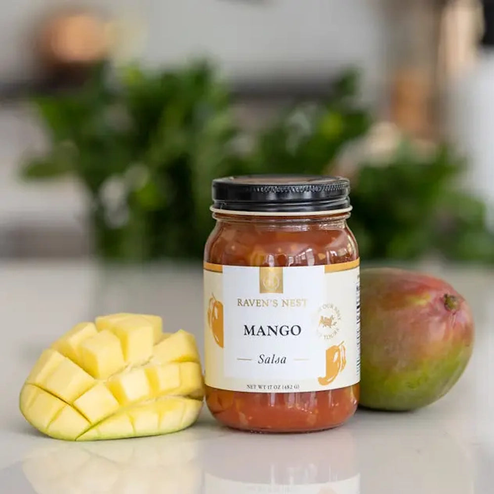 A glass jar of Raven’s Nest Mango Salsa on a white counter next to a whole mango and a sliced mango half.