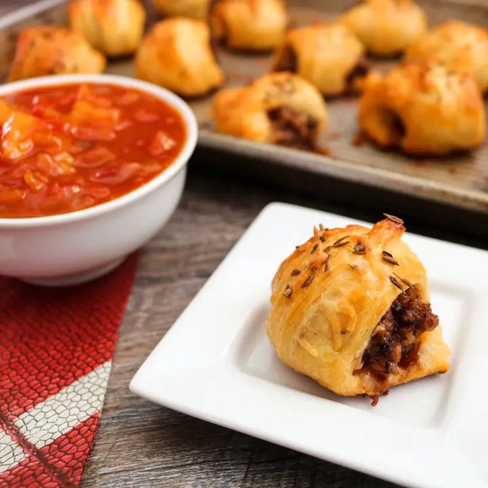 Golden pastry puff filled with meat on a small white plate, with a bowl of red salsa in the background.
