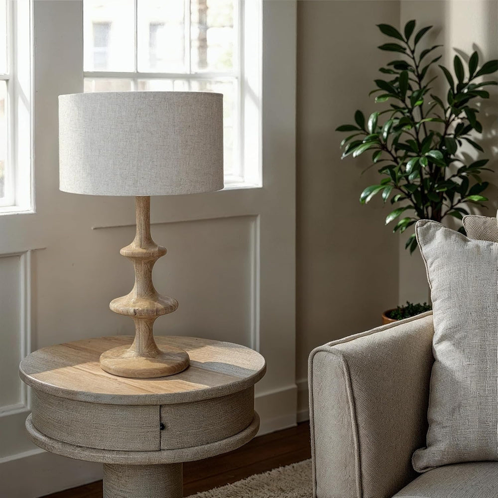 Sculptural light wood table lamp with linen shade on a round pedestal side table in a bright, modern living room.