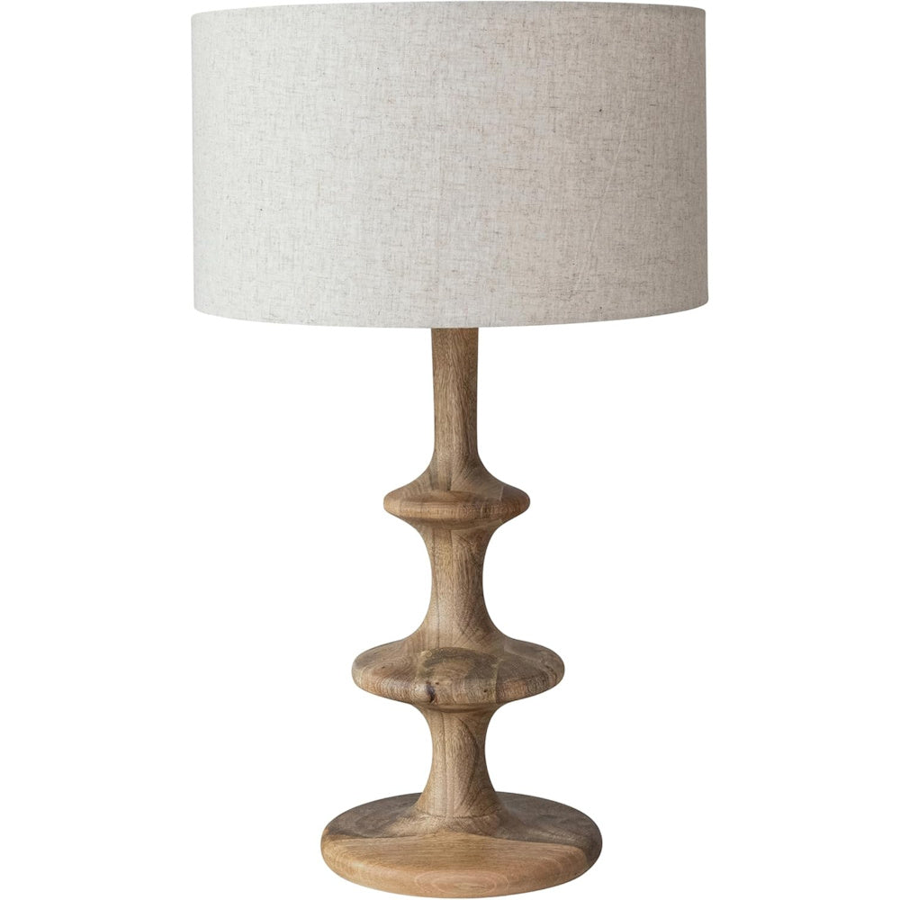 Carved wooden table lamp with a tiered spindle base and off-white linen drum shade for rustic home decor.