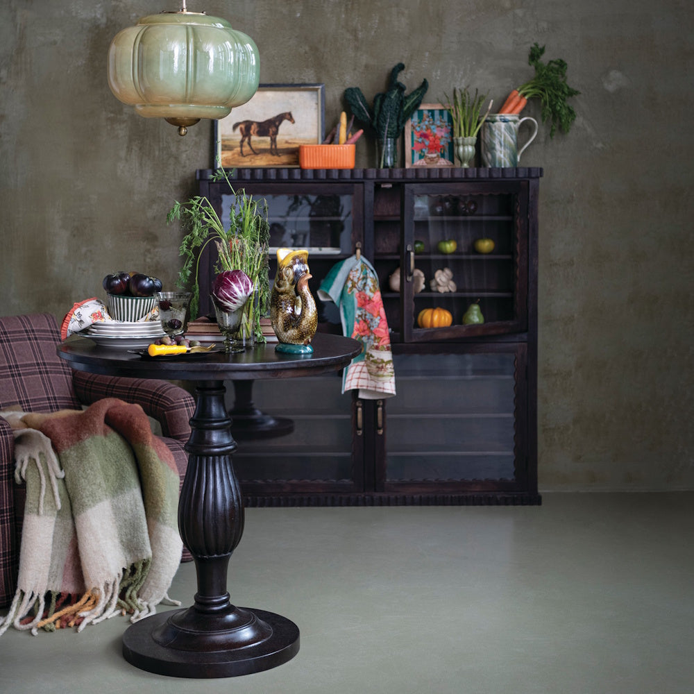 Round dark mango wood pedestal table with a fluted turned base in a rustic dining room setting.