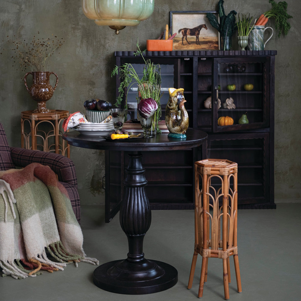 Round dark mango wood pedestal dining table with a carved turned base in a rustic, moody dining room setting.