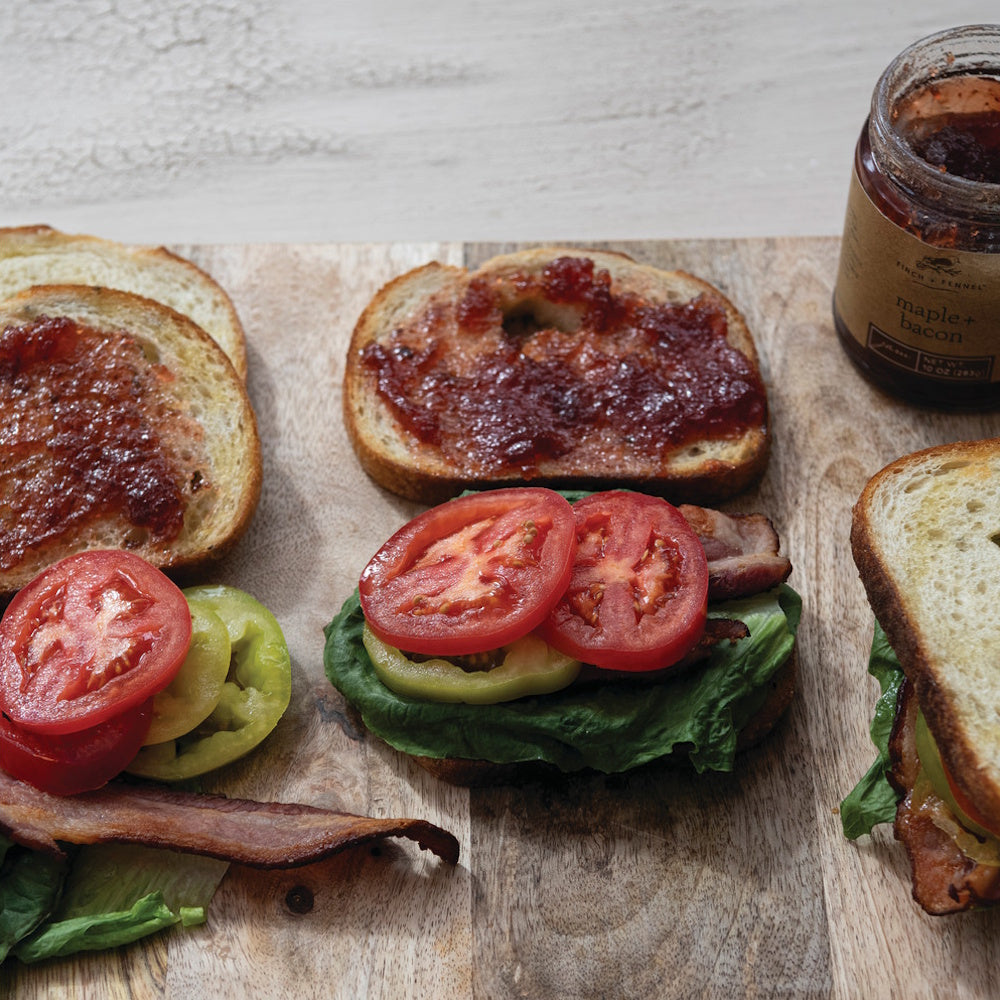 Sandwiches being prepared with lettuce, tomatoes, bacon, and Finch + Fennel maple bacon jam on a cutting board.