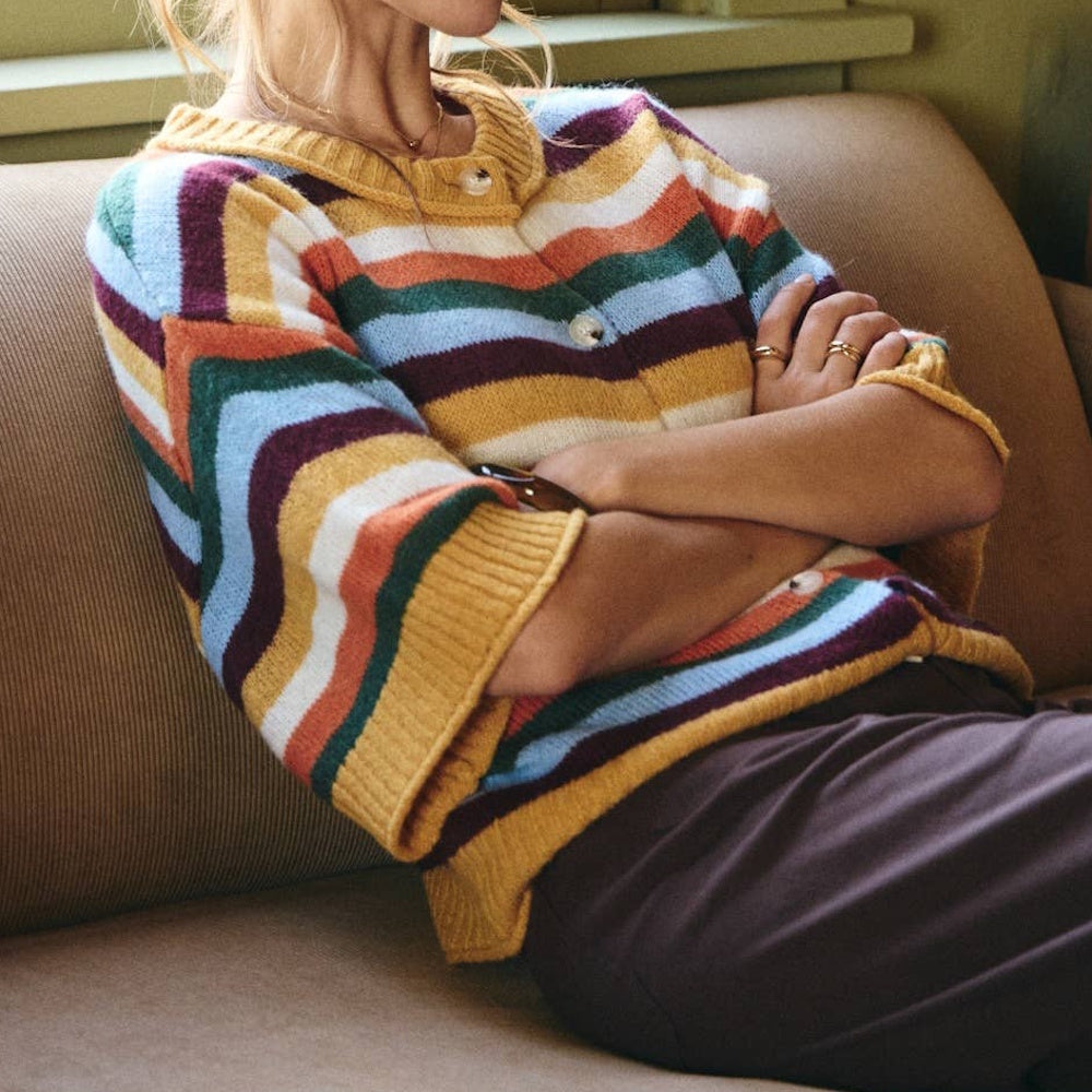 Woman sitting on sofa wearing a multi-color striped knit short-sleeve cardigan sweater with front buttons.