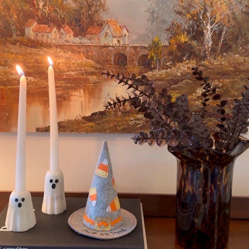 Two white ghost taper candle holders with lit candles and a felt witch hat on display.