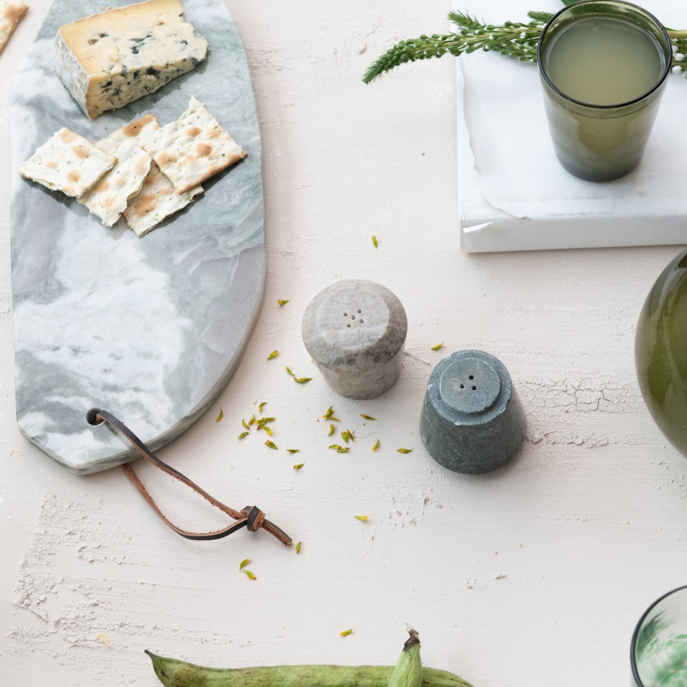 Two marble salt and pepper shakers next to a green marble serving board with cheese and crackers.