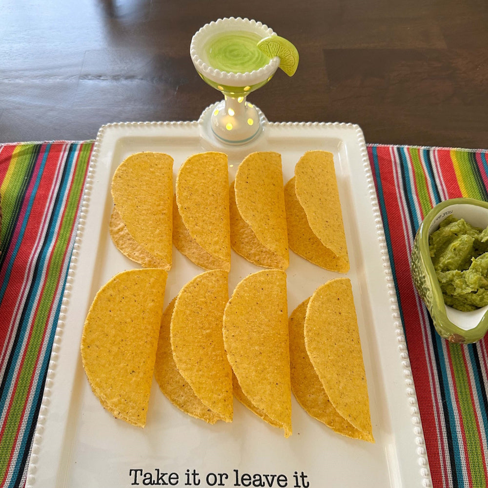 Eight hard taco shells on a white platter with guacamole and a decorative margarita light on a colorful striped tablecloth.