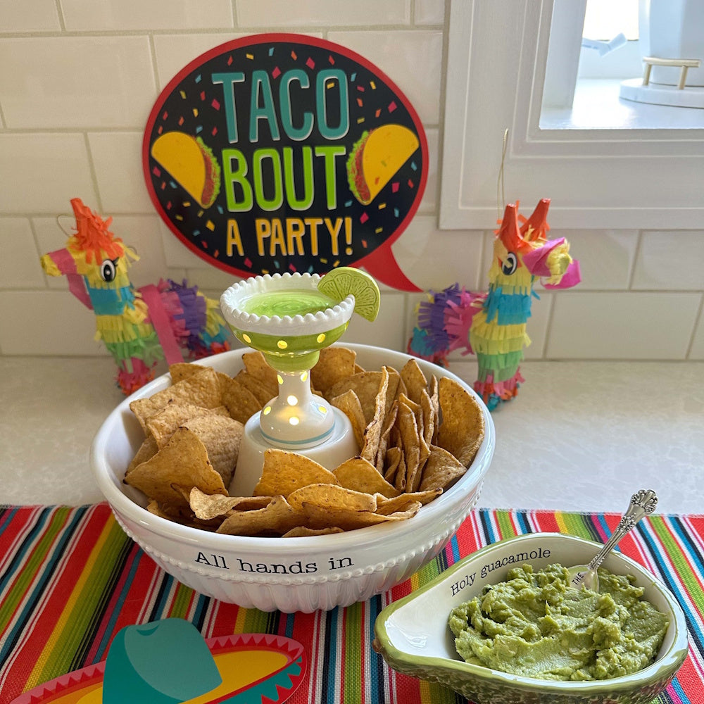 Festive taco party spread with chips, guacamole, and a light-up margarita sitter centerpiece on a colorful striped runner.
