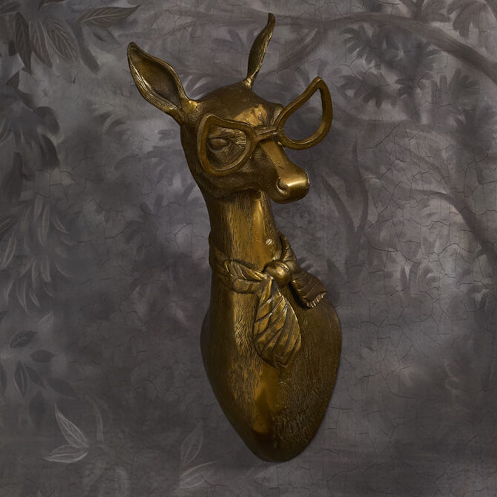 A brass-finished deer head wall mount wearing quirky cat-eye glasses and a knotted neck scarf against a dark background.