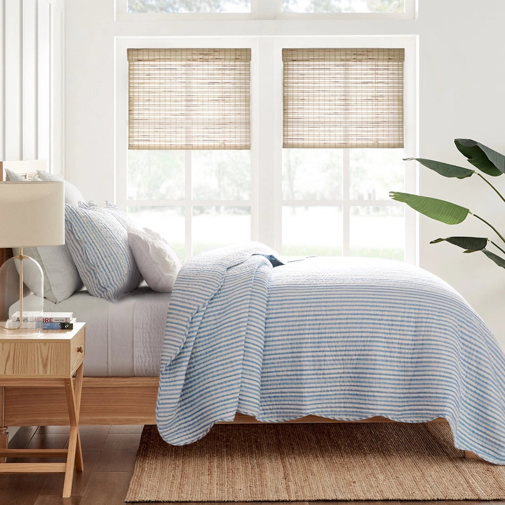 Side view of a bed dressed in a blue and white striped quilt set with matching shams in a bright room.
