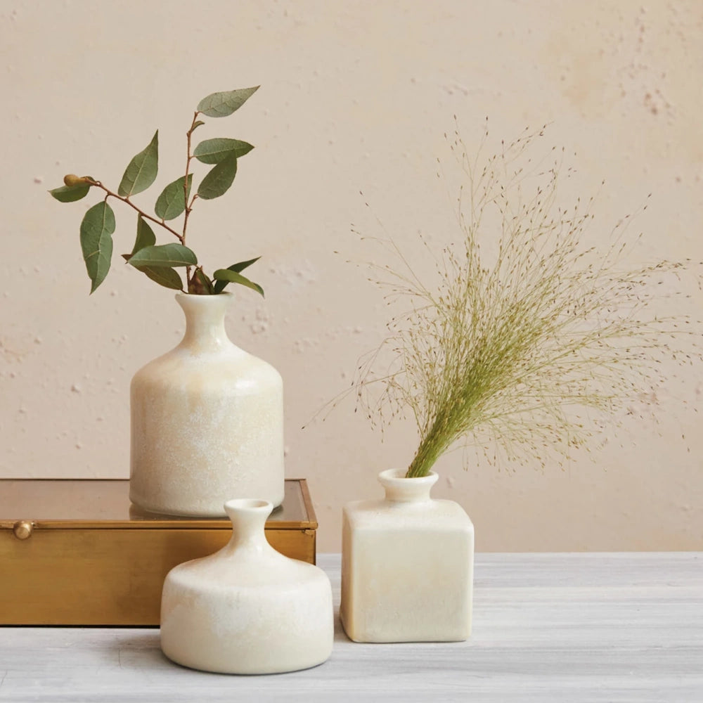 Three small off-white ceramic vases with foliage arranged on a surface next to a decorative gold glass box.
