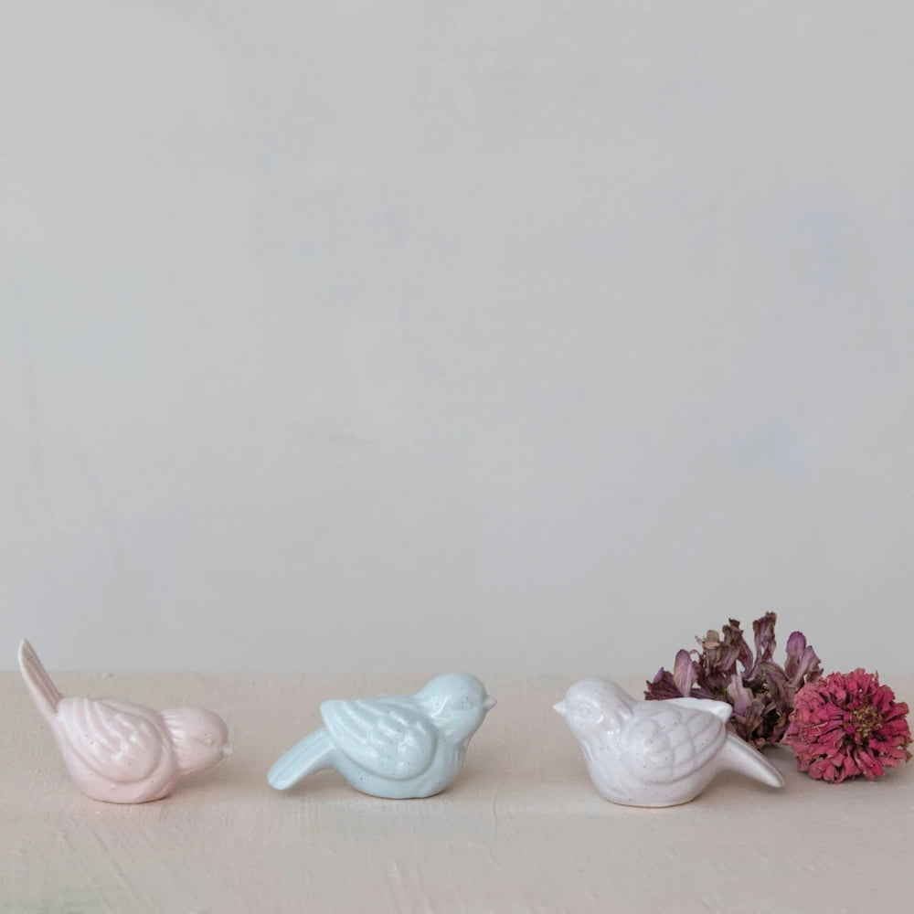 Three small matte-finish stoneware bird figurines in pink, blue, and white next to dried flowers.