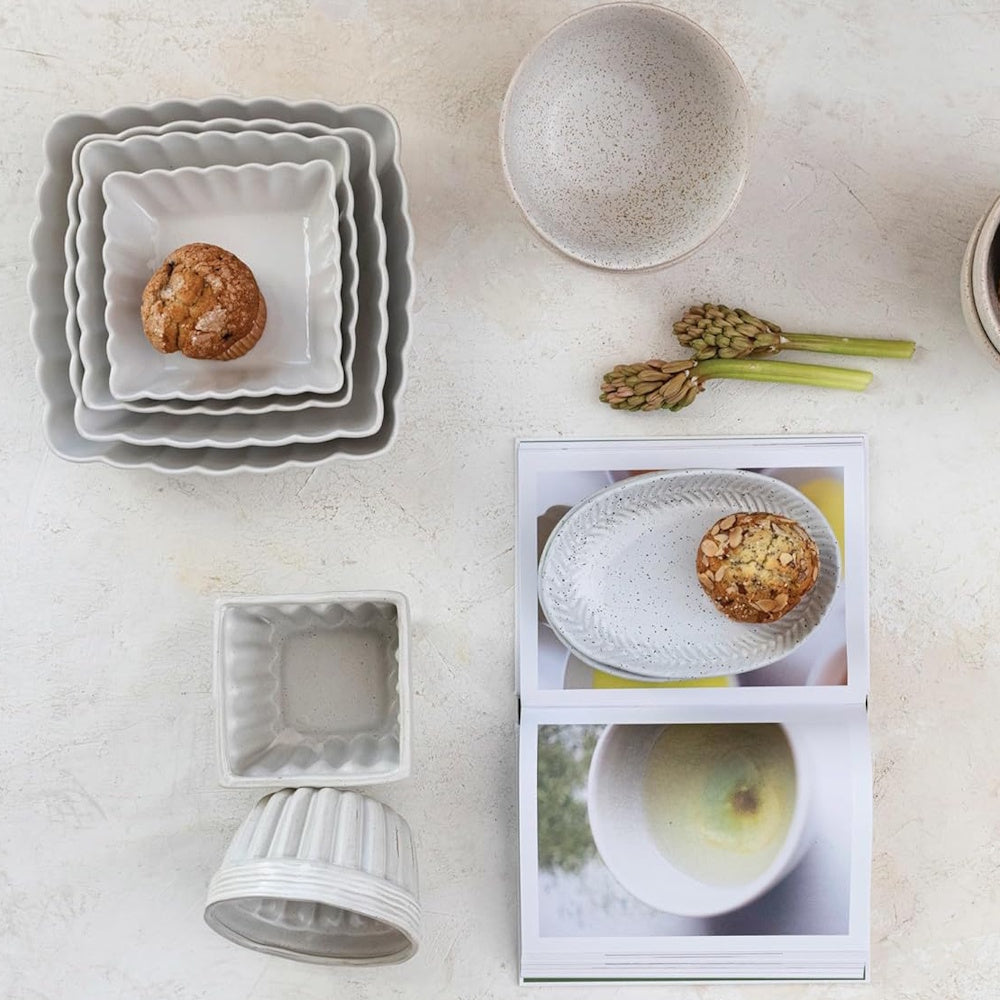 Flat lay of scalloped nesting bowls, a muffin, speckled stoneware, and an open cookbook on a light table.