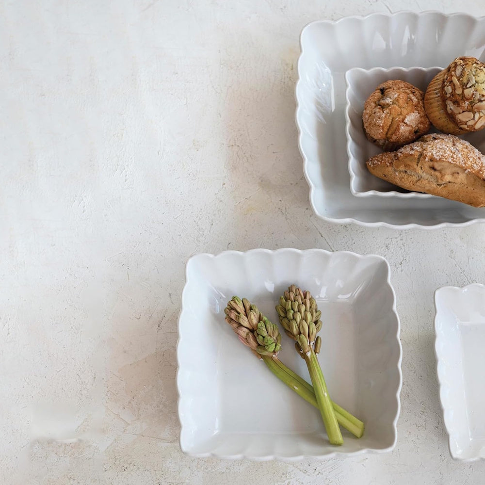 White square stoneware bowls with ruffled edges holding pastries and flower buds on a textured tabletop.