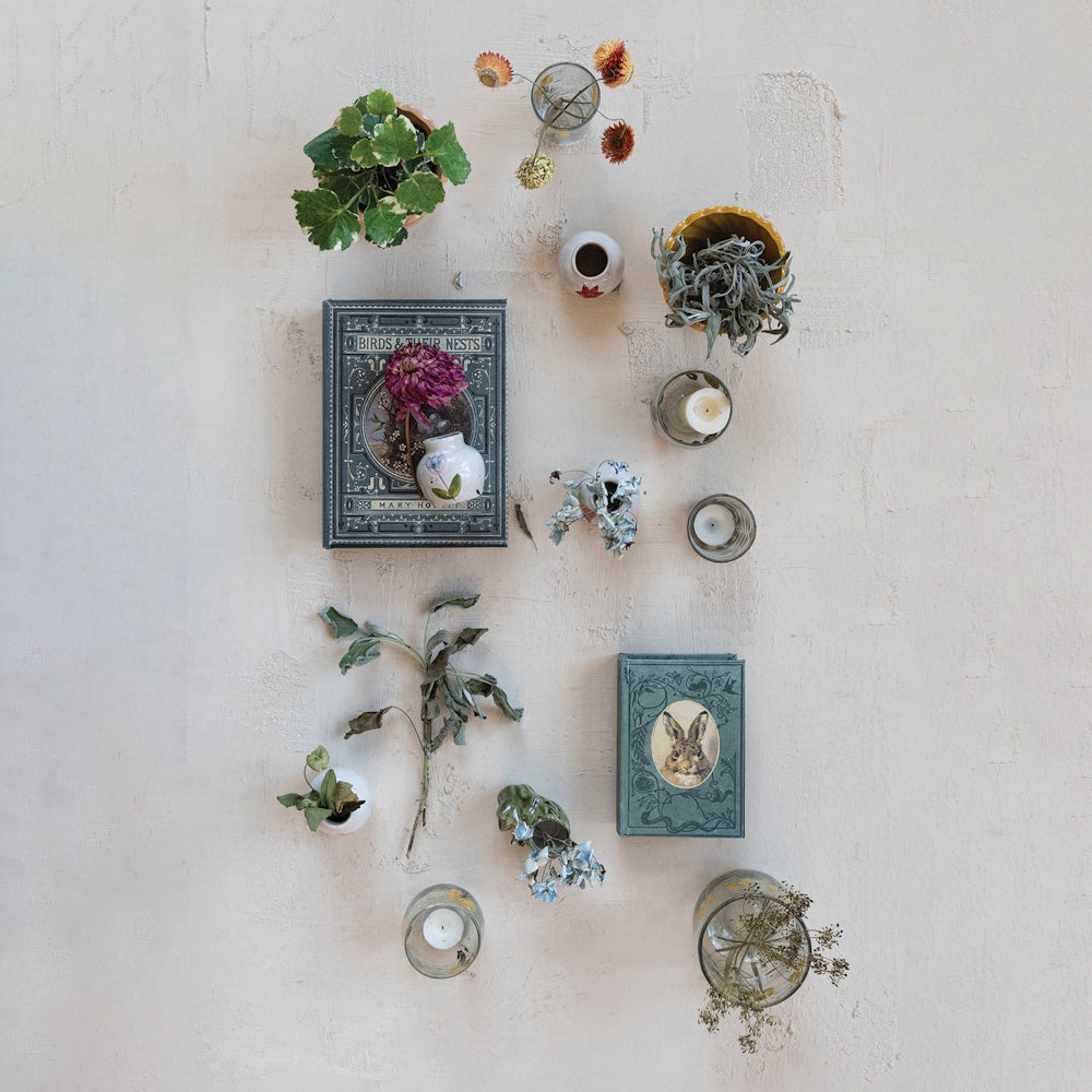 Flat lay of vintage book-shaped boxes, potted plants, dried flowers, and small vases on a textured surface.