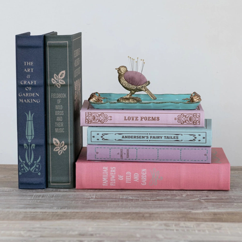 Vintage style decorative book boxes stacked with a gold bird pin cushion on a turquoise tray.