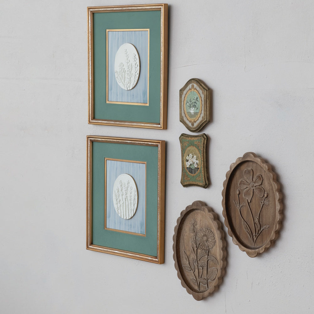 Lifestyle side view of a botanical gallery wall arrangement featuring framed art and carved wooden plaques.