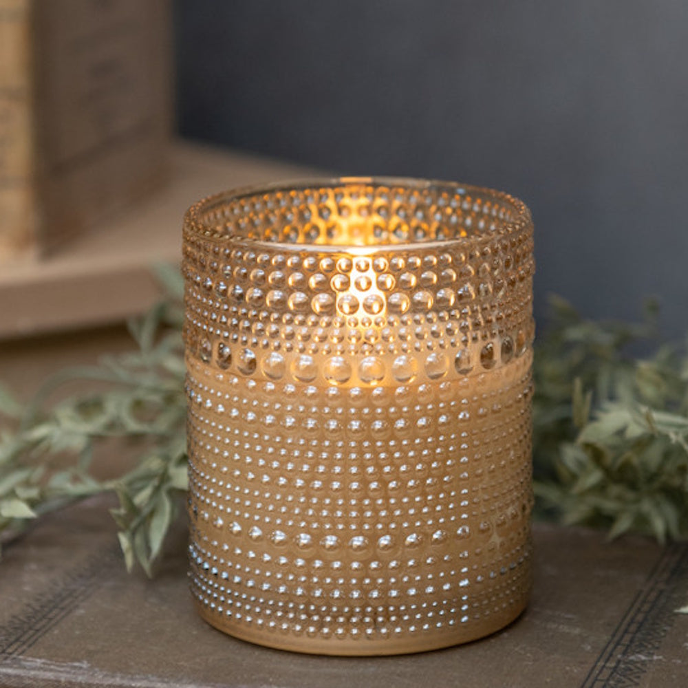 Lit amber dotted glass candle holder casting a warm glow on a wooden surface with greenery behind.