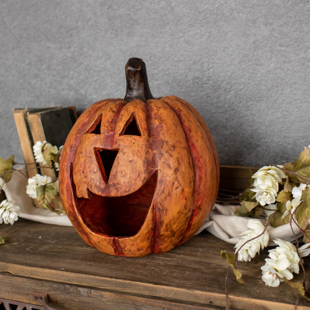 Carved rustic orange jack-o'-lantern with a wide mouth resting on a shelf with white flowers and old books.