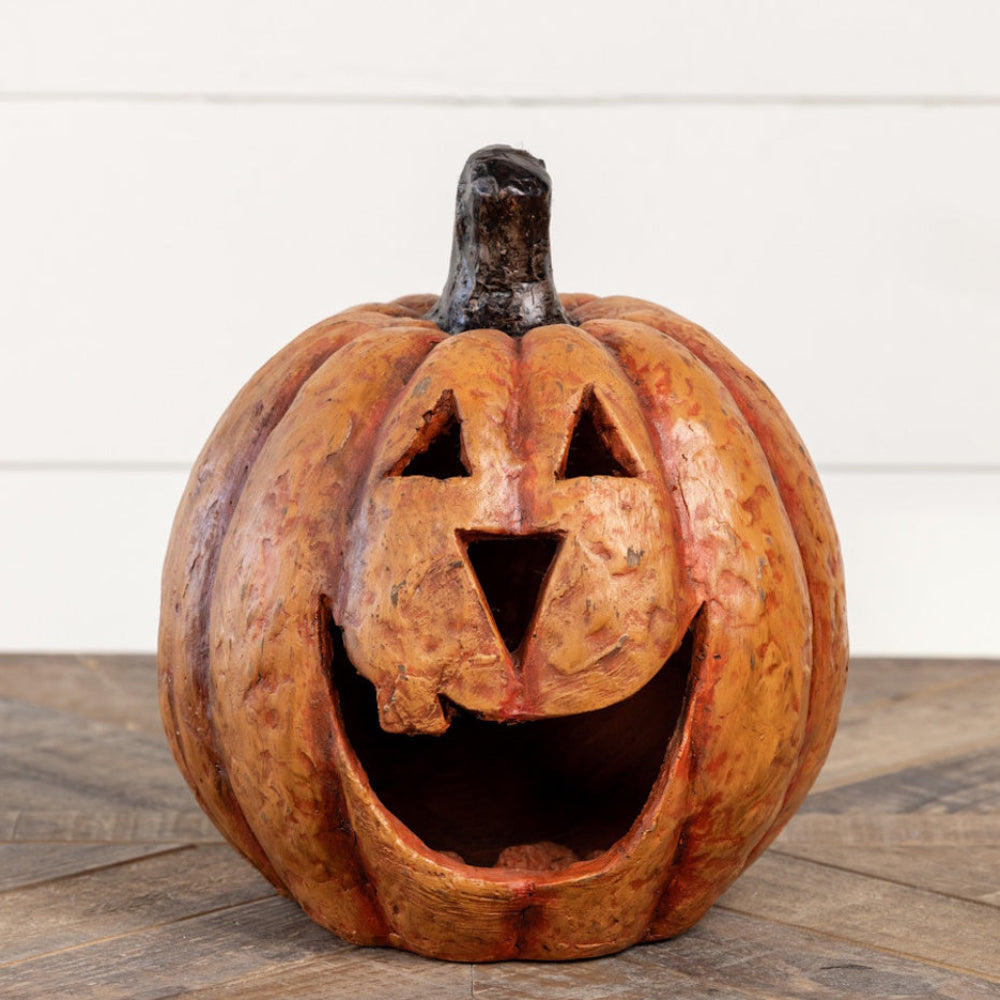 Carved rustic orange jack-o'-lantern with a wide smiling mouth on a wooden surface against a white wall.