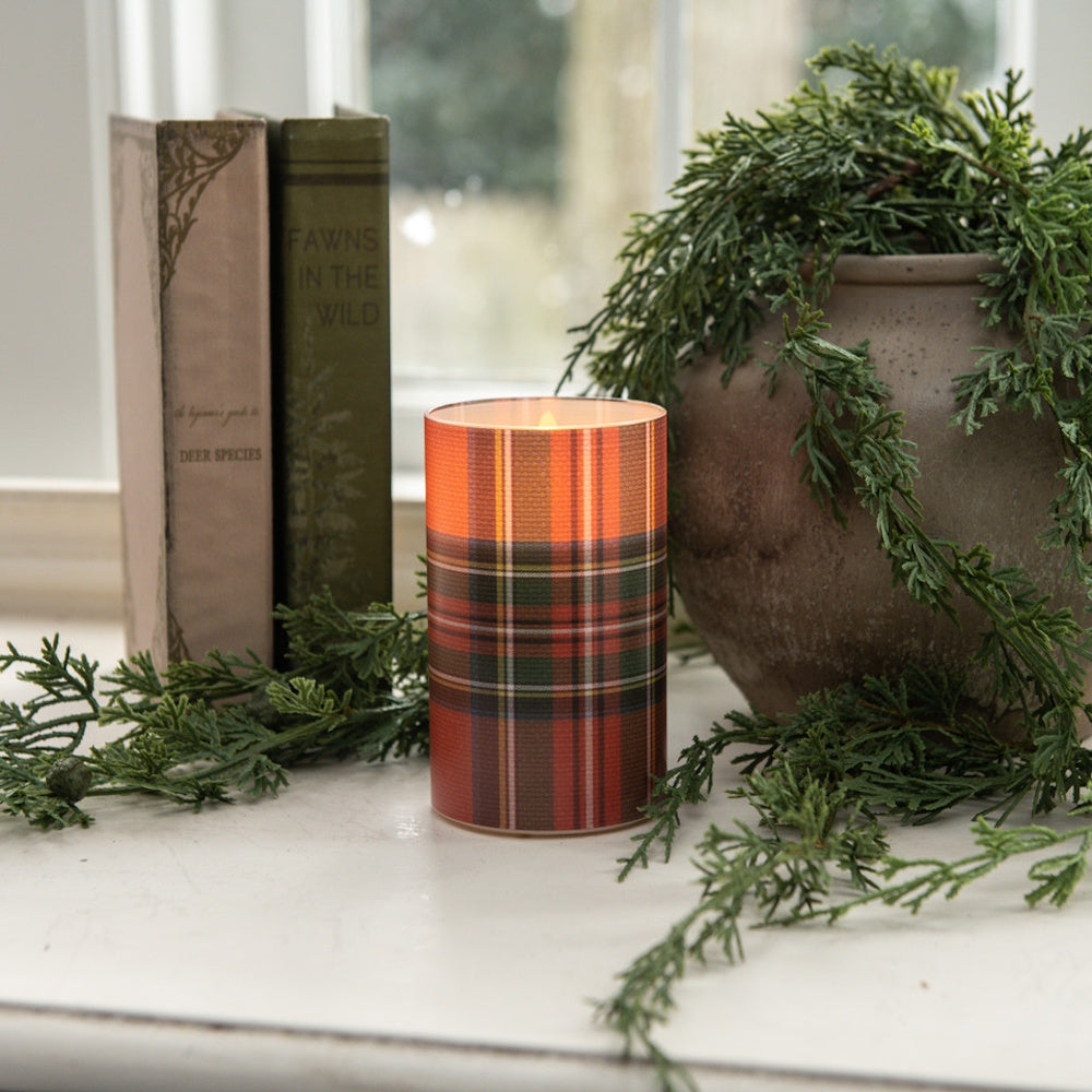 Glowing red tartan plaid candle on a white windowsill next to vintage books and greenery.