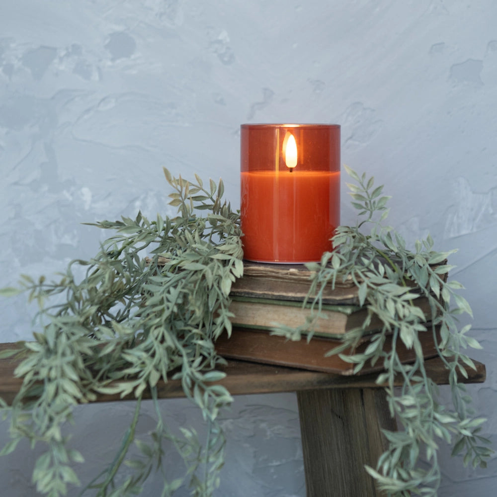 Lit sienna-orange glass candle on stacked books with cascading green garland.